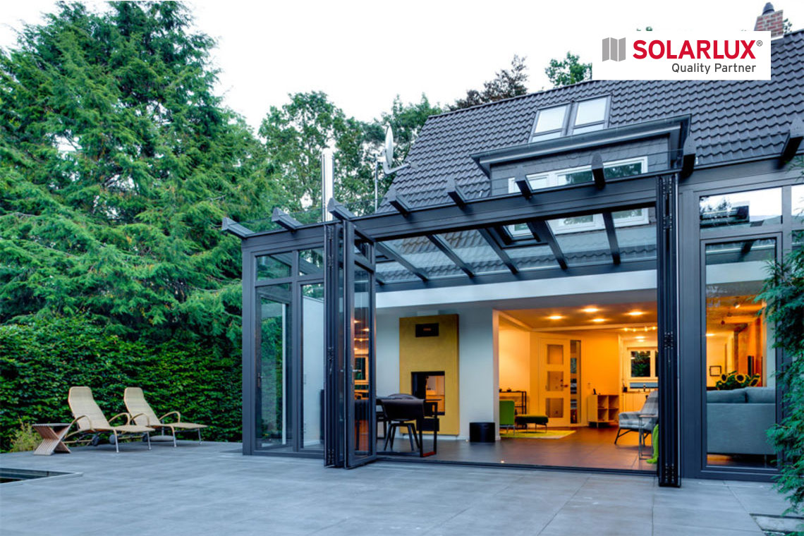Blick von der Terrasse auf einen Wintergarten mit gegenläufigem Dach, die Glasfaltwand zur Terrasse hin ist geöffnet, das Innere des Hauses ist wohnlich beleuchtet.
